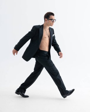 Smokinli dinamik genç adamın yan görünüşü. Tişörtü yok, yana bakıyor ve açık gri arkaplan önünde büyük adımlarla hızla yürüyor.