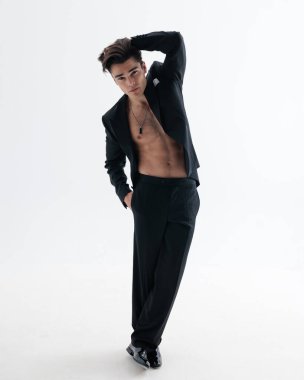 Smokinli şehvetli, zarif bir adam karın kaslarını gösteriyor, elleri başının arkasında, açık gri arka planda el ele duruyor.