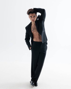 Şehvetli genç iş adamı bir eli başının arkasında diğeri cebinde açık gri arka planda gömleksiz bir smokin giyerken poz veriyor.