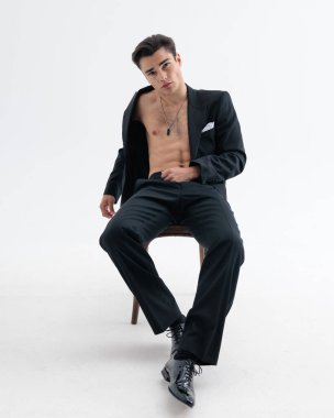 Seksi, zarif iş adamı smokinsiz bir şekilde sandalyede oturuyor ve açık gri arka planda havalı bir şekilde poz veriyor.