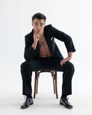 Smokinli, gömleksiz, çenesine dokunan ve açık gri arka planda tahta koltukta otururken düşünen zarif bir adam.