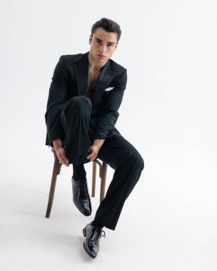 Smokinli, gömleksiz havalı moda işadamı açık gri arka planda oturup beklerken çoraplarını düzeltiyor.