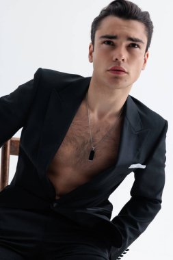 Siyah smokinli çekici genç bir işadamı, açık gri arka planda otururken göğsünü gösteriyor.