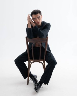 Siyah smokinli şehvetli genç adam, açık gri arka planda tahta bir sandalyede otururken, çıplak göğsünü kapalı ellere yaslıyor.