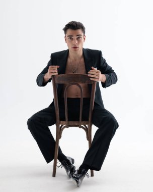 Açık siyah smokin giyen kendine güvenen genç adam göğsünü tahta bir sandalyede geriye doğru oturtur ve açık gri arka planda ileriye bakar.