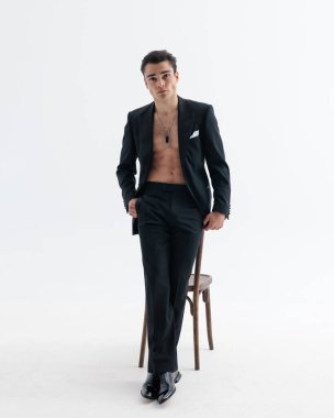 Smokinli kendine güvenen iş adamı, çıplak göğsü ceplerinde el ele tutuşmuş, öne bakıyor ve açık gri arka planda sandalyeye yaslanıyor.