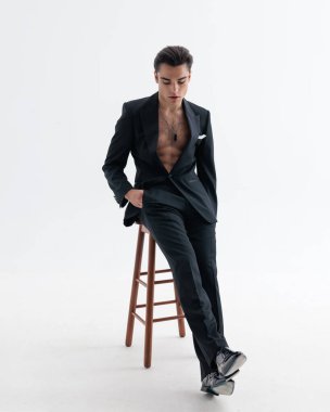 Açık smokin giyen, aşağı bakarken karın kaslarını ve göğsünü gösteren, el ele tutuşan ve açık gri arka planda sandalyeye yaslanan zarif fit bir adam.