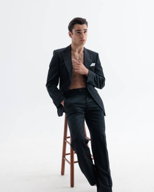 Siyah smokinli, gömleksiz seksi bir genç adam el ele poz verirken diğer elini açık gri arka planda göğsüne kaldırdı.