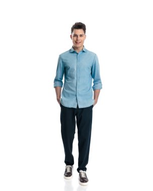 Mavi kot gömlekli mutlu genç adam elleri ceplerinde beyaz arka planda hareket ediyor.