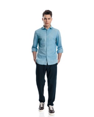 Beyaz arka planda elleri ceplerinde, kot gömlekli rahat bir adam yürüyor.