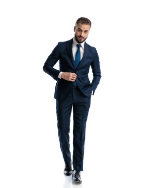 Seksi, zarif adam lacivert takımı ayarlıyor ve beyaz arka planda güvenle yürüyor.