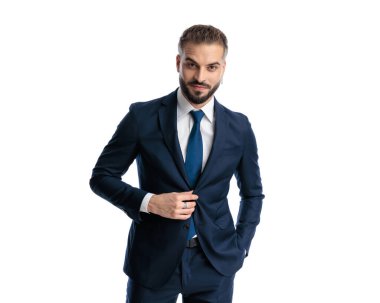 Yakışıklı, olgun bir adam ileriye bakıyor, gülümsüyor ve beyaz arka planda dururken lacivert takımını ayarlıyor.