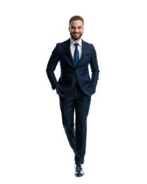 Lacivert takımlı yakışıklı bir adam gülümsüyor ve elleri ceplerinde beyaz arka planda yürüyor.