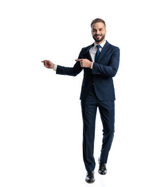Heyecanlı, lacivert takım elbiseli zarif genç adam, parmaklarını sağa sola gösteriyor, gülümsüyor ve beyaz arka planda yürüyor.