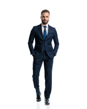 Lacivert takım elbiseli çekici bir adam seksi ve elleri ceplerinde beyaz arka planda yürüyor.