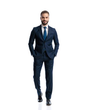 Lacivert takım elbiseli mutlu bir işadamı beyaz arka planda elleri ceplerinde yürüyor.