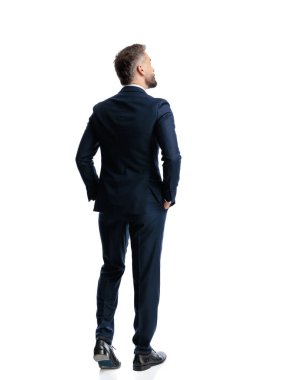 Lacivert takım elbiseli zarif olgun bir adamın beyaz arka planda elleri ceplerinde yürümesi.