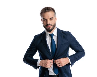 Kendine güvenen olgun bir adamın portresi. Beyaz arka planda dikilirken lacivert takımının düğmelerini açıyor.