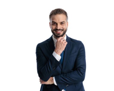 Deniz mavisi takımlı mutlu iş adamının portresi. Çeneye dokunuyor, gülümsüyor ve beyaz arka planda poz verirken düşünüyor.