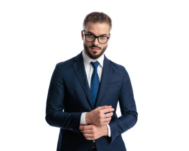 Gözlüklü yakışıklı olgun bir adam lacivert takım elbise kollarını ayarlıyor ve beyaz arka planda poz veriyor.