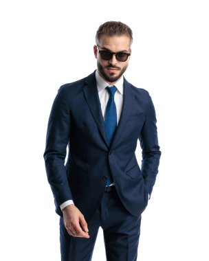 Lacivert takım elbiseli, güneş gözlüklü, elini cebine atıp beyaz arka planda duran seksi, olgun bir adam.