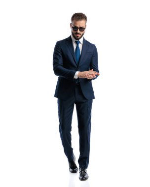Moda özgüveni olan lacivert takım elbiseli, güneş gözlüklü, avuçlarını beyaz arka planda gezdiren bir adam. 