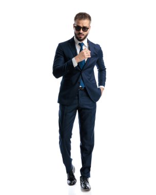 Lacivert takım elbiseli şık bir iş adamı beyaz arka planda elleri ceplerinde yürüyor.