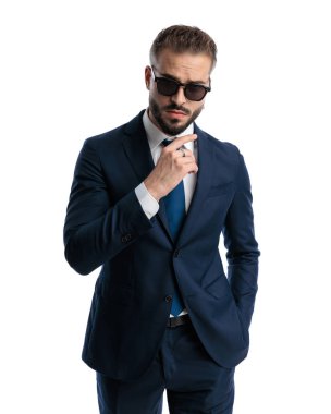 Güneş gözlüklü, lacivert elbiseli, beyaz arka planda elleri ceplerinde poz veren havalı moda adamı portresi.