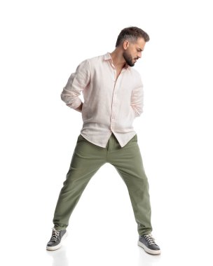 Havalı bir genç adam beyaz arka planda dikilirken arkasını kollayıp poz veriyor.