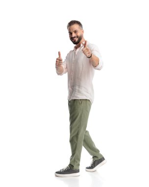 Mutlu genç adam gülümsüyor ve beyaz arka planda yürürken başparmak işareti yapıyor.