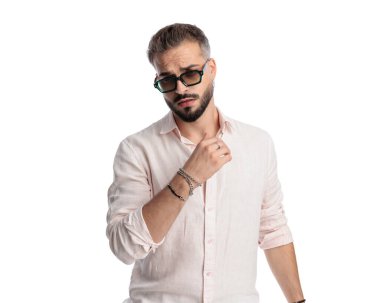 Sakallı, güneş gözlüklü, beyaz arka planda havalı poz veren genç bir adam.