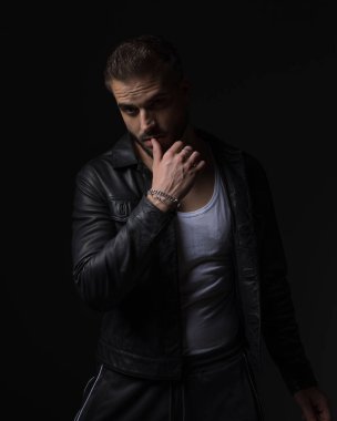 Siyah deri ceketli, baştan çıkarıcı bir gencin portresi dudaklara dokunuyor ve siyah arka planda poz veriyor.