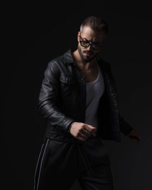 Deri ceketli sakallı genç adam aşağı bakıyor ve siyah arka planda havalı bir poz veriyor.