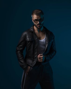 Kendine güveni tam, güneş gözlüklü ve mavi arka planda siyah deri ceket giyerken öne bakıyor.