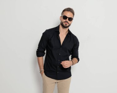 Güneş gözlüklü, el ele tutuşan ve gri arka planda poz veren kendinden emin bir gencin portresi.