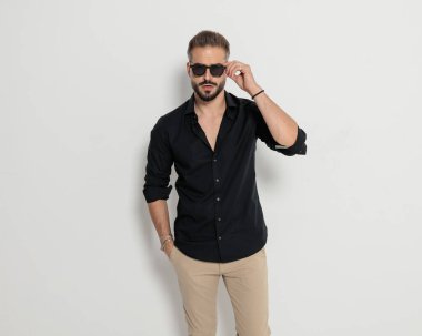 Gri arka planda havalı bir şekilde poz verirken el ele tutuşup güneş gözlüğü takan çekici, zeki ve sıradan bir adamın portresi.