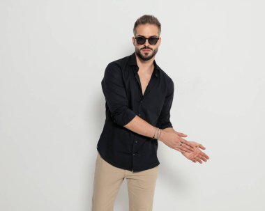 Güneş gözlüklü seksi sakallı adam avuç içlerine sürtünüyor, poz veriyor ve gri arka planda duruyor.