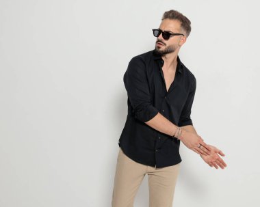 Siyah gözlüklü, avuçlarını yana doğru ovuşturan ve gri arka planda diğer tarafa bakan havalı genç adam.