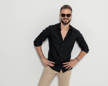 Güneş gözlüklü, kalçasında el ele tutuşan ve gri arka planda gülümseyen mutlu, zeki bir adam.