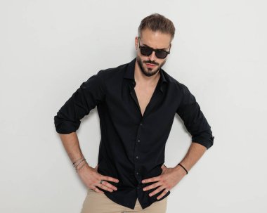 Siyah gözlüklü kibirli genç adam kalça üzerinde el ele tutuşuyor ve gri arka planda duruyor.