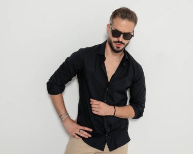 Güneş gözlüklü, kalçasında el ele tutuşan ve gri arka planda poz veren havalı moda adamı portresi.