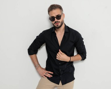 Şehvetli, güneş gözlüklü, siyah gömlekli ve elleri kalçasında gri arka planda poz veren genç bir adam.