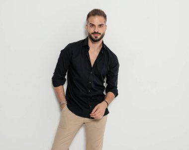 Siyah gömlekli mutlu bir adamın portresi. Ceplerinde el ele tutuşmuş, gülümsüyor ve gri arka planda poz veriyor.