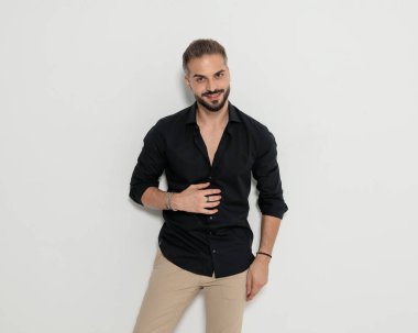 Siyah gömlekli çekici genç adam gri arka planda dururken ileri bakıyor ve gülümsüyor.