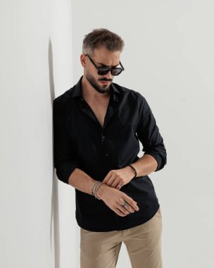 Güneş gözlüklü yakışıklı genç adam aşağı bakıyor, bilezikleri tamir ediyor ve gri bir duvara yaslanıyor.