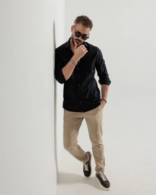 Güneş gözlüklü, şehvetli genç adam dudaklarına dokunuyor, el ele tutuşuyor ve gri arka planda bir duvara yaslanıyor.