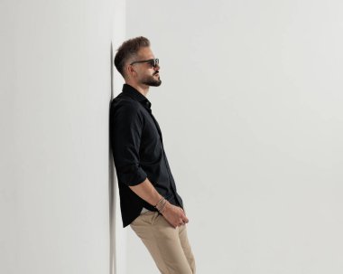 Güneş gözlüklü, gri duvara yaslanmış, yan gözle bakan havalı bir genç adam. 