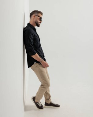 Güneş gözlüklü sakallı genç adam duvara yaslanıyor ve gri arkaplanın önüne bakıyor.