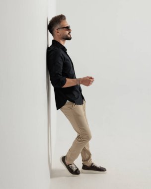 Güneş gözlüklü seksi sakallı adam yana bakıyor ve gri arka planda bir duvara yaslanıyor.