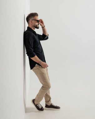 Yandan bakan seksi genç adamın yan görünüşü, güneş gözlüklerini ayarlamak ve gri arka planın önündeki duvara yaslanmak.